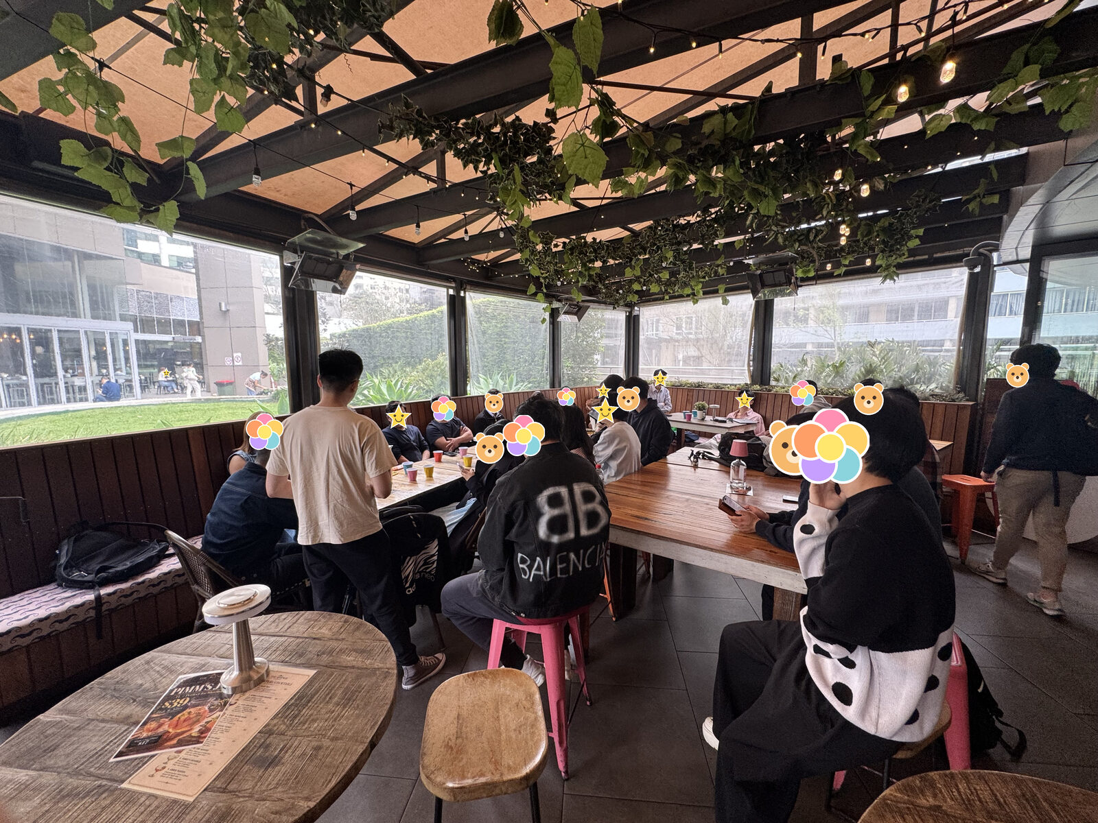 玻璃房的全景，头顶是藤，长桌两边坐满了人，前景是一张圆木桌